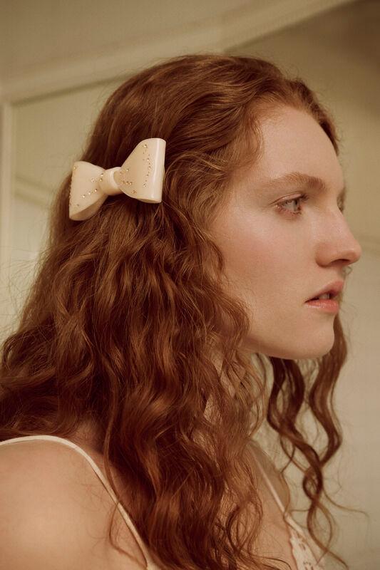 Maanesten Ribin Hair Clip Cream zeigt eine cremefarbene Haarspange in Schleifenform auf langem lockigem Haar. Modell 3981