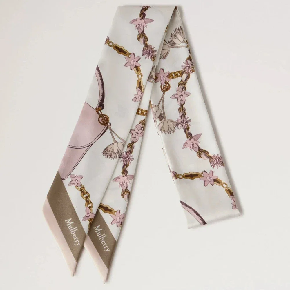 Mulberry Lana Bag and Chains Skinny Scarve Eggshell/Blossom Pink präsentiert einen schmalen Seidenschal mit rosa und goldenen Kettendetails. Modell VS4849115W569