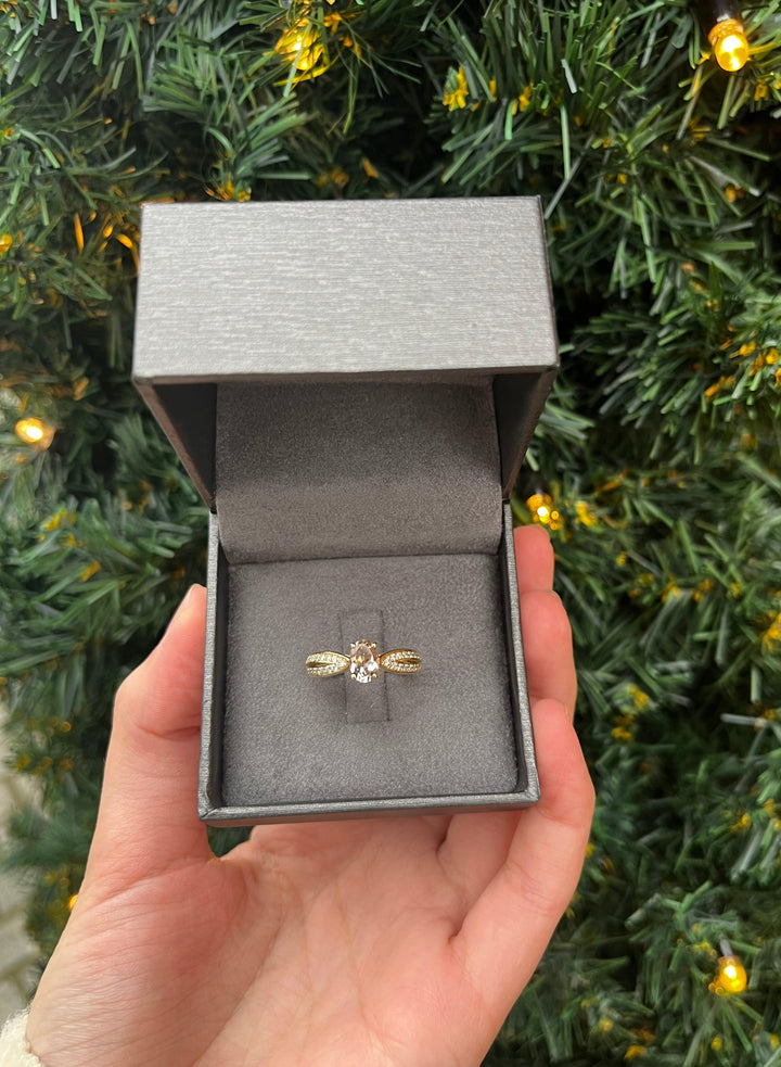 Nuran 14kt Silvia Ring 0,30ct WSI präsentiert in grauer Box, goldenes Design vor dem Weihnachtsbaum. Modell a2455-mo-030-rg-001