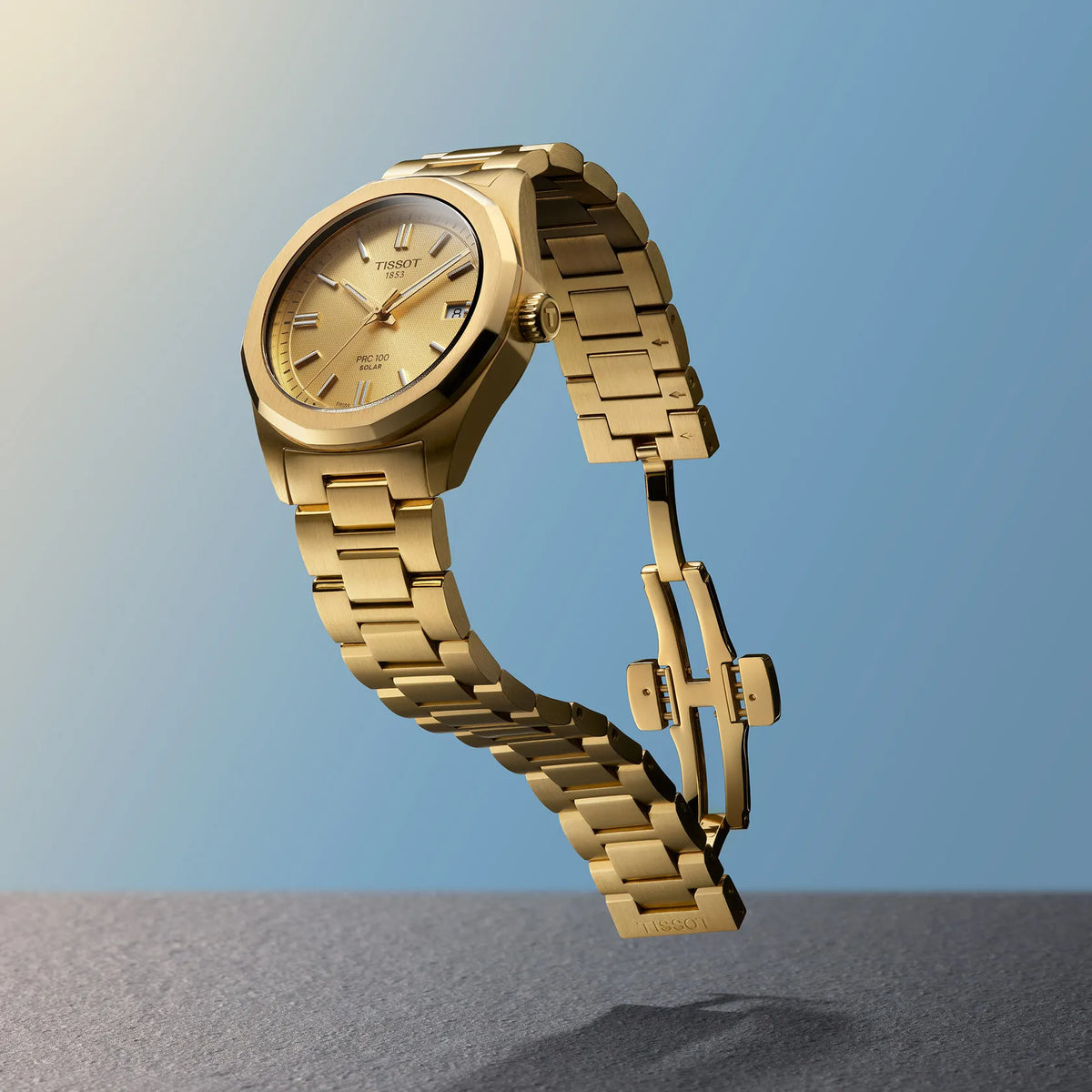 Tissot PRC 100 Solar 34 mm Gold-PVD-Uhr mit goldenem Zifferblatt und Stahlkette vor blauem Hintergrund. Modell T1518223302100