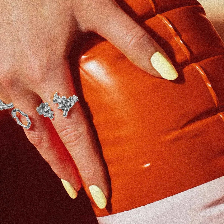 Vincent Orb Chronicles Solv Ring, eleganter Silberring mit organischem Design auf der Hand mit hellen Nägeln vor orangefarbenem Hintergrund. Modell VJ163-URS-001-2