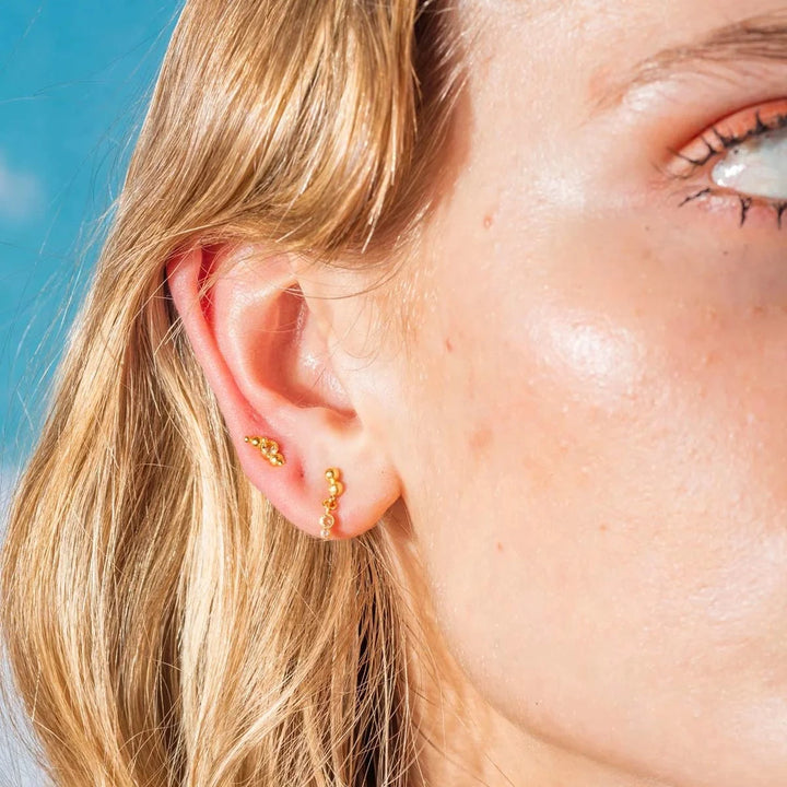 Vincent Pendelohrringe, vergoldet, am Ohr abgebildet, kleine Kugeln in Goldoptik unter klarem Himmel. Modell VJ177-UEG-2