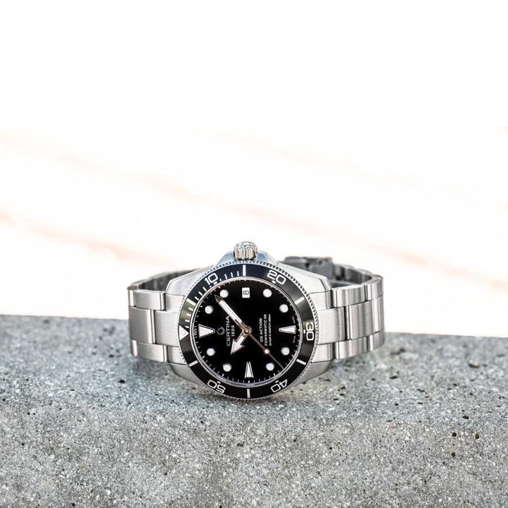 Certina DS Action Powermatic 80 Herrenuhr mit schwarzem Zifferblatt und Stahlkette auf grauem Beton, Modell C0328071105100.