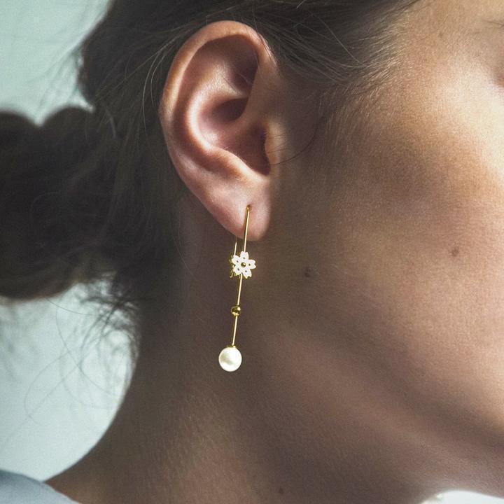 Am Ohr ist ein vergoldeter Ohrring mit hängender Perle von Polar Jewelry zu sehen, mit einer goldenen Blume und einer Perle. Modell SAK-EH-GD-WP-00120